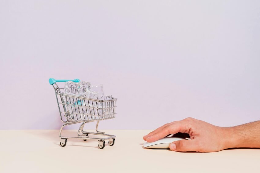 Conceptual image of a hand on a mouse next to a miniature shopping cart filled with ice cubes, symbolizing online shopping.